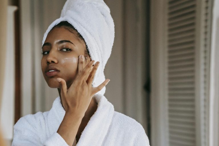 Young ethnic female in robe with terry towel on head applying moisturizing cream on face while looking in mirror in house